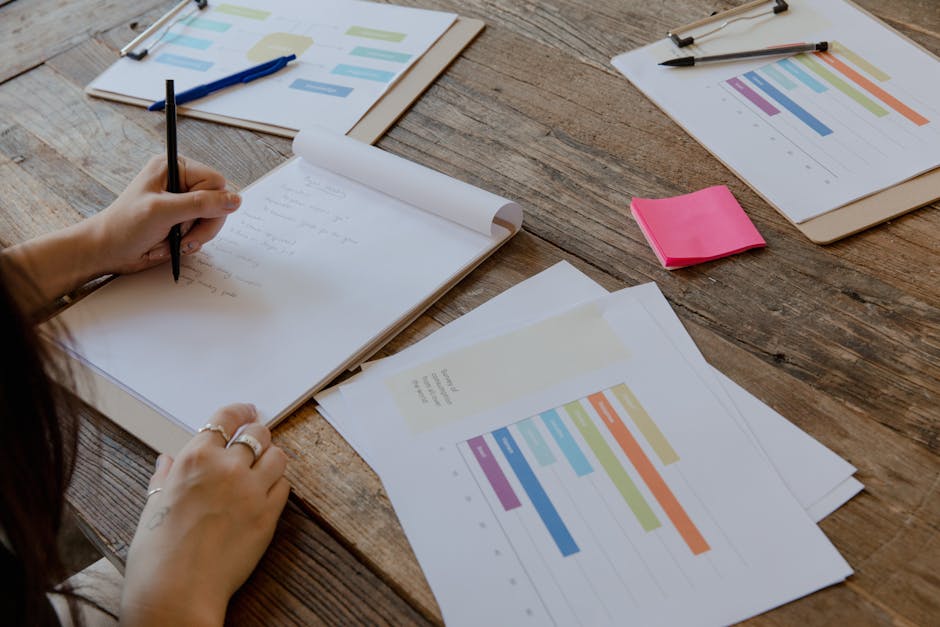 Hands writing on paper with colorful charts on wooden table, organized workspace.